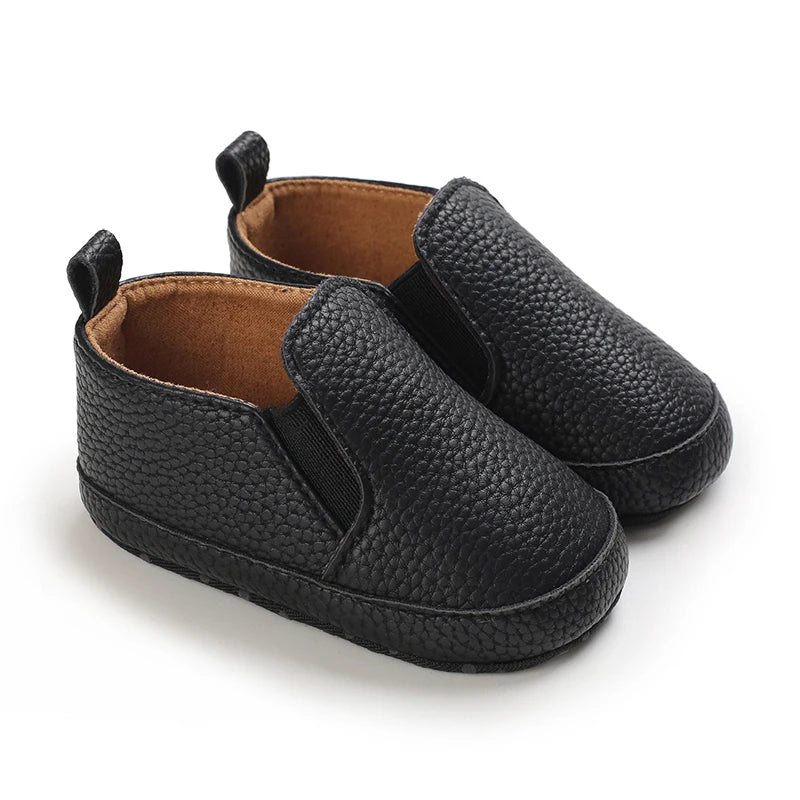 A pair of black faux leather baby first walker sneakers with a dinosaur print detail.
