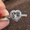 A close-up of a heart-shaped cubic zirconia diamond ring being held by a person against a dark brown background.