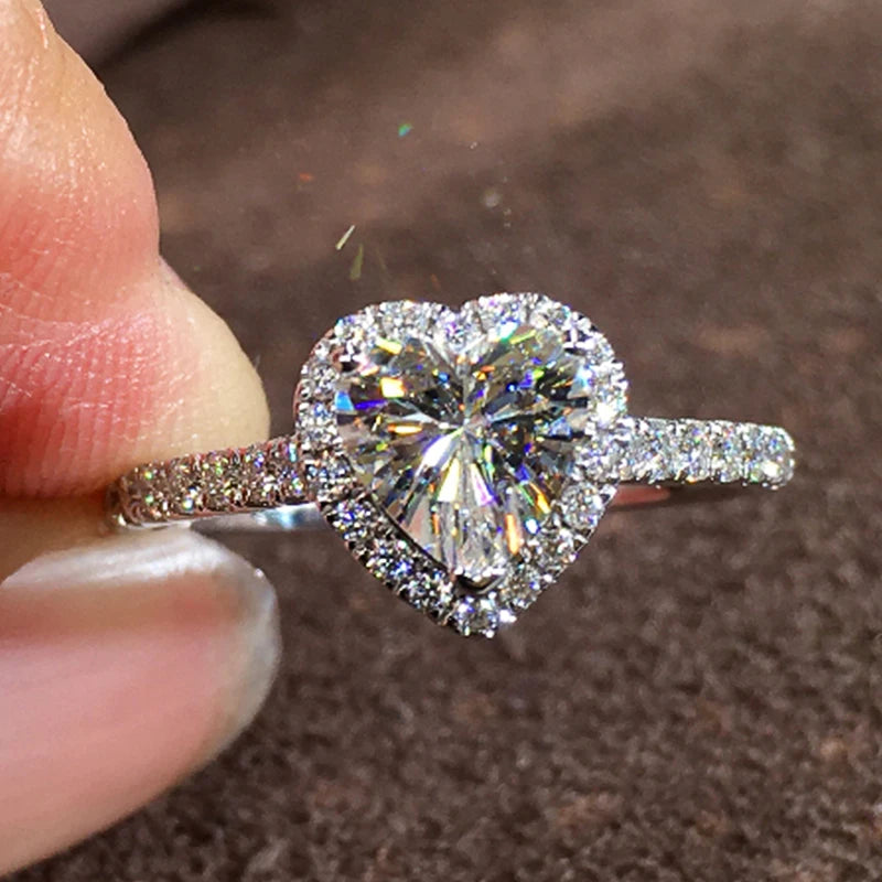 A close-up of a heart-shaped cubic zirconia diamond ring being held by a person against a dark brown background.
