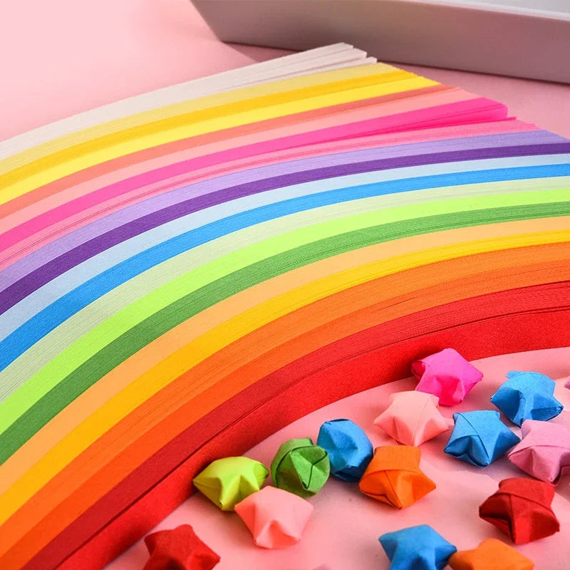 Colorful origami star strips in a rainbow gradient are laid out on pink paper for crafting