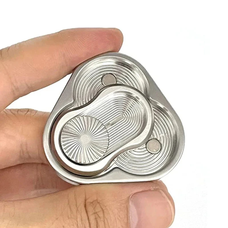 A hand holds a silver metal magnetic fidget slider with concentric grooves and small spheres.