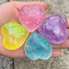 A hand holds three heart-shaped squishy toys in pink, purple, and blue glitter foam.