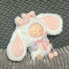 A white rabbit costume for a doll featuring pink ears and a bow, set against a striped background.