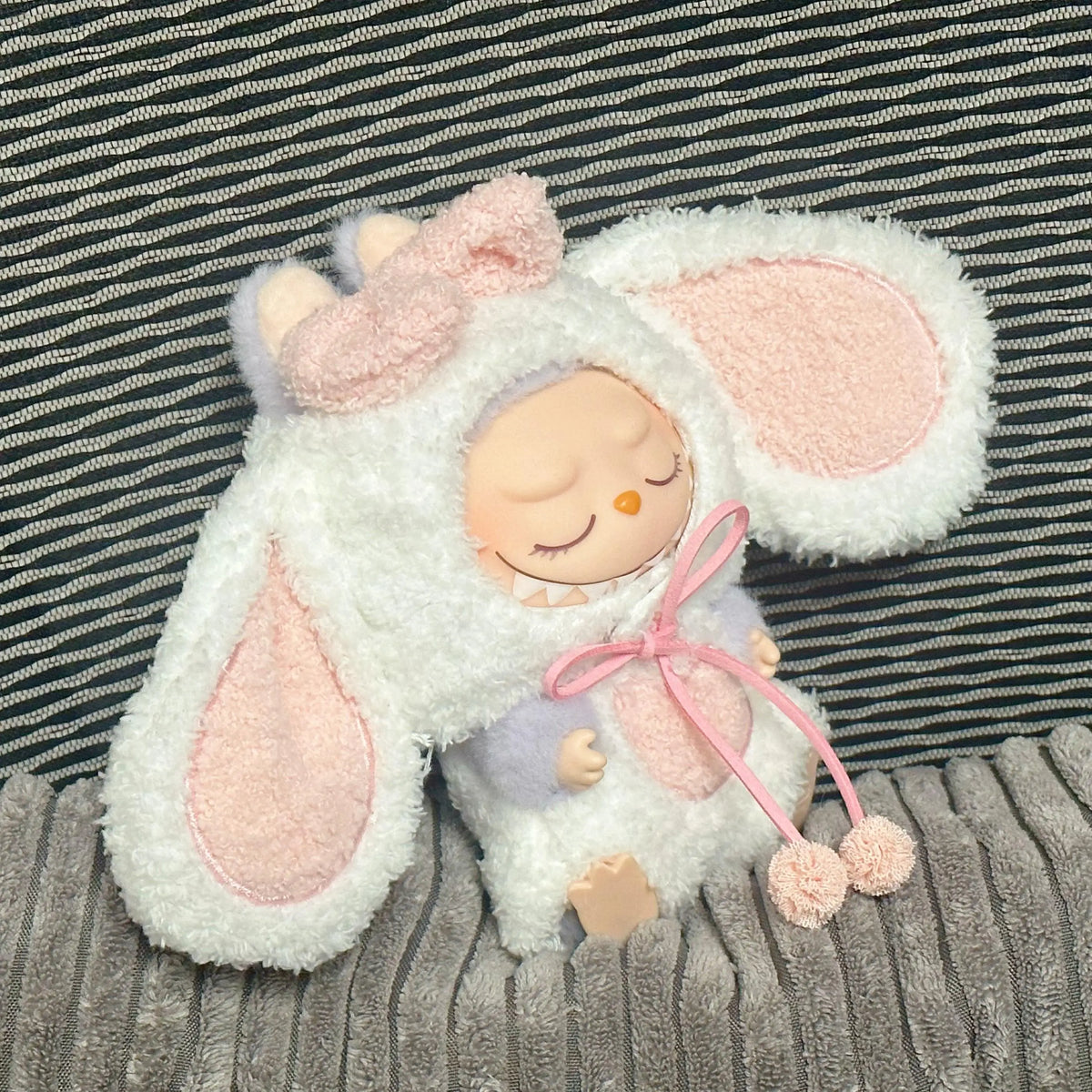 A white rabbit costume for a doll featuring pink ears and a bow, set against a striped background.