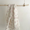 A white cotton muslin swaddle blanket with a pink floral print is hanging on a light wooden dowel.