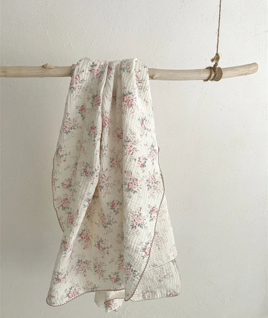 A white cotton muslin swaddle blanket with a pink floral print is hanging on a light wooden dowel.
