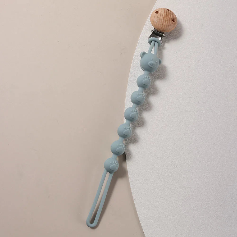 A light blue silicone bear paw pacifier clip with a wooden top and a flexible silicone tube.
