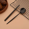 A black stainless steel Korean dinnerware set including chopsticks and a spoon placed on a black wire rack.