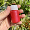 A hand holds a red metallic gold acrylic paint bottle with a white cap against a blurred green outdoor background.