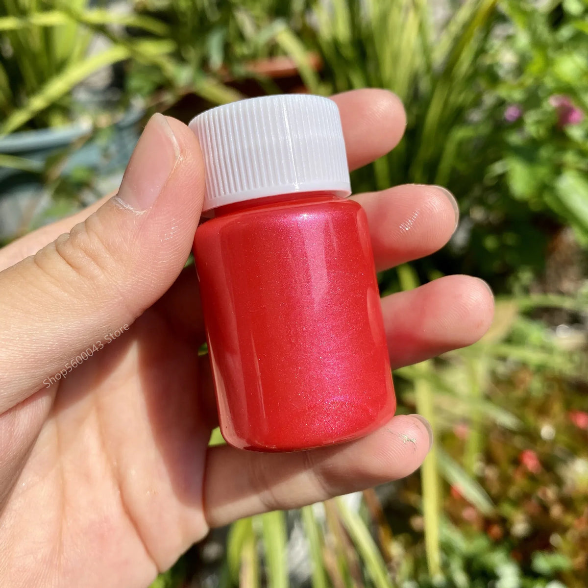 A hand holds a red metallic gold acrylic paint bottle with a white cap against a blurred green outdoor background.