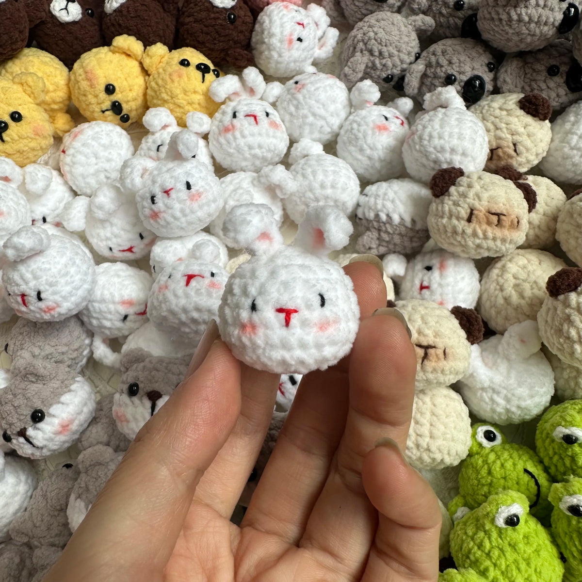 A hand holds a small white crocheted animal keychain, possibly a rabbit or chick, among many similar handmade items.