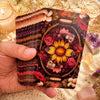 A hand holds a tarot deck featuring floral and earthy tones, set against a gold textured background.