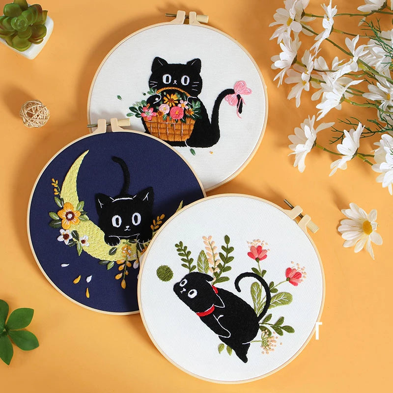 Three embroidery kits featuring black cats and floral designs on white fabric are displayed on a light orange background.