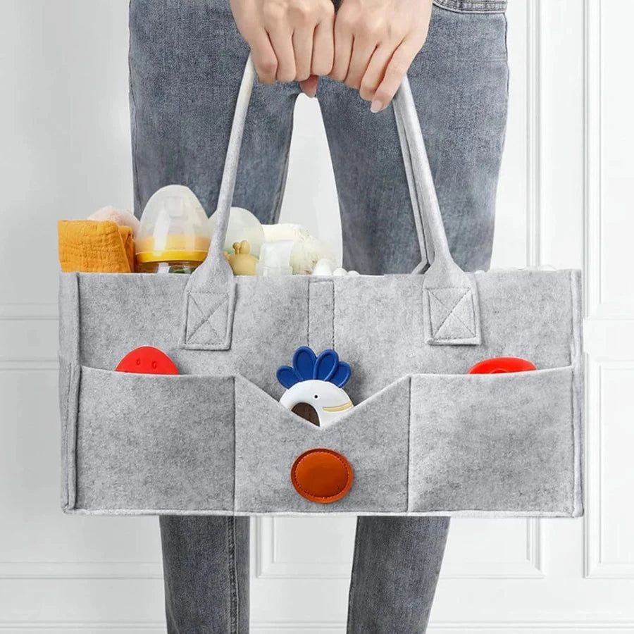 A gray felt diaper storage bag with multiple compartments and colorful baby items is held by a person.