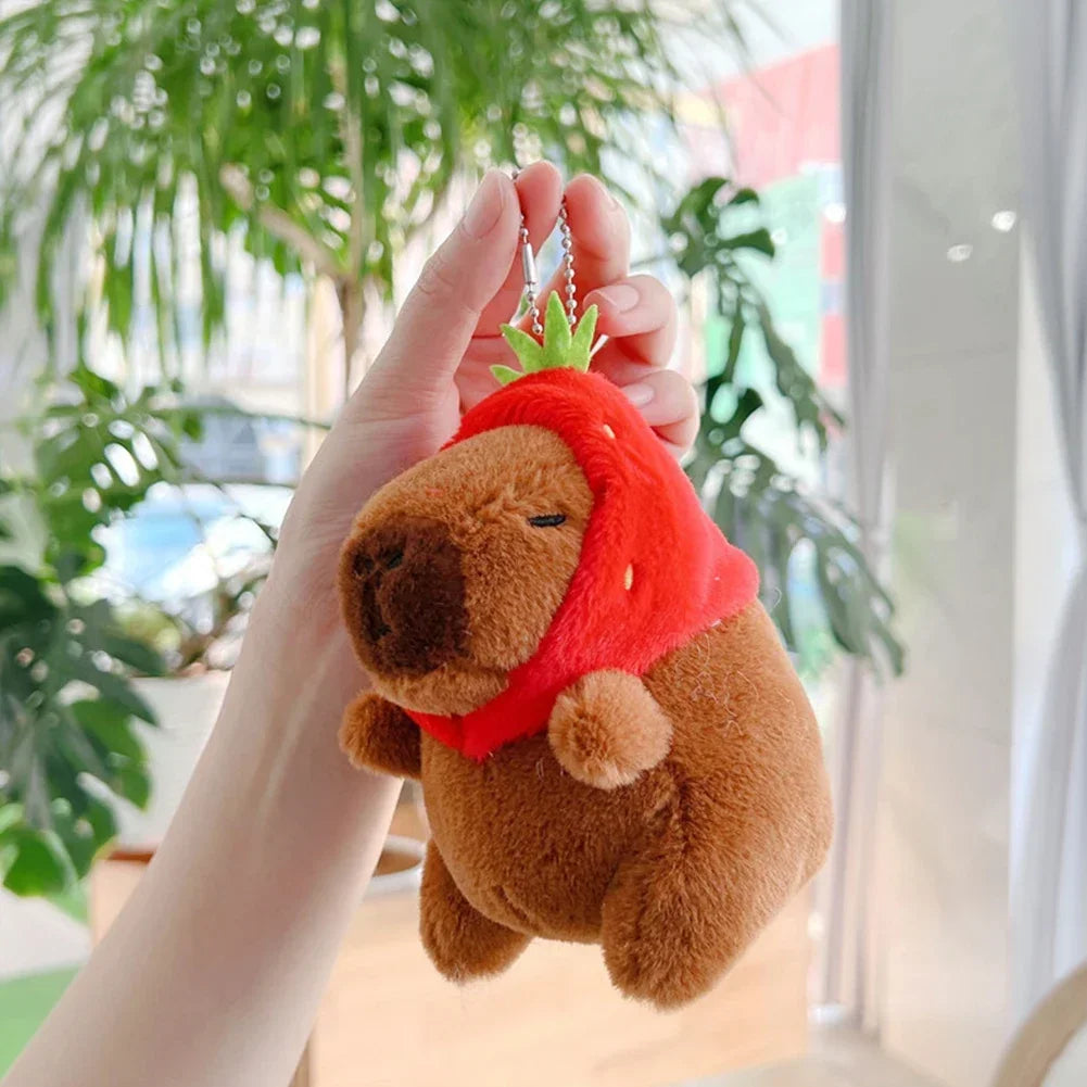 A brown capybara plush toy wearing a red bandana is held by a hand, displayed as a keychain charm.