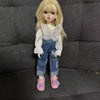 A fashion doll wearing denim overalls and a white shirt with a butterfly detail, standing in front of a dark gray couch.