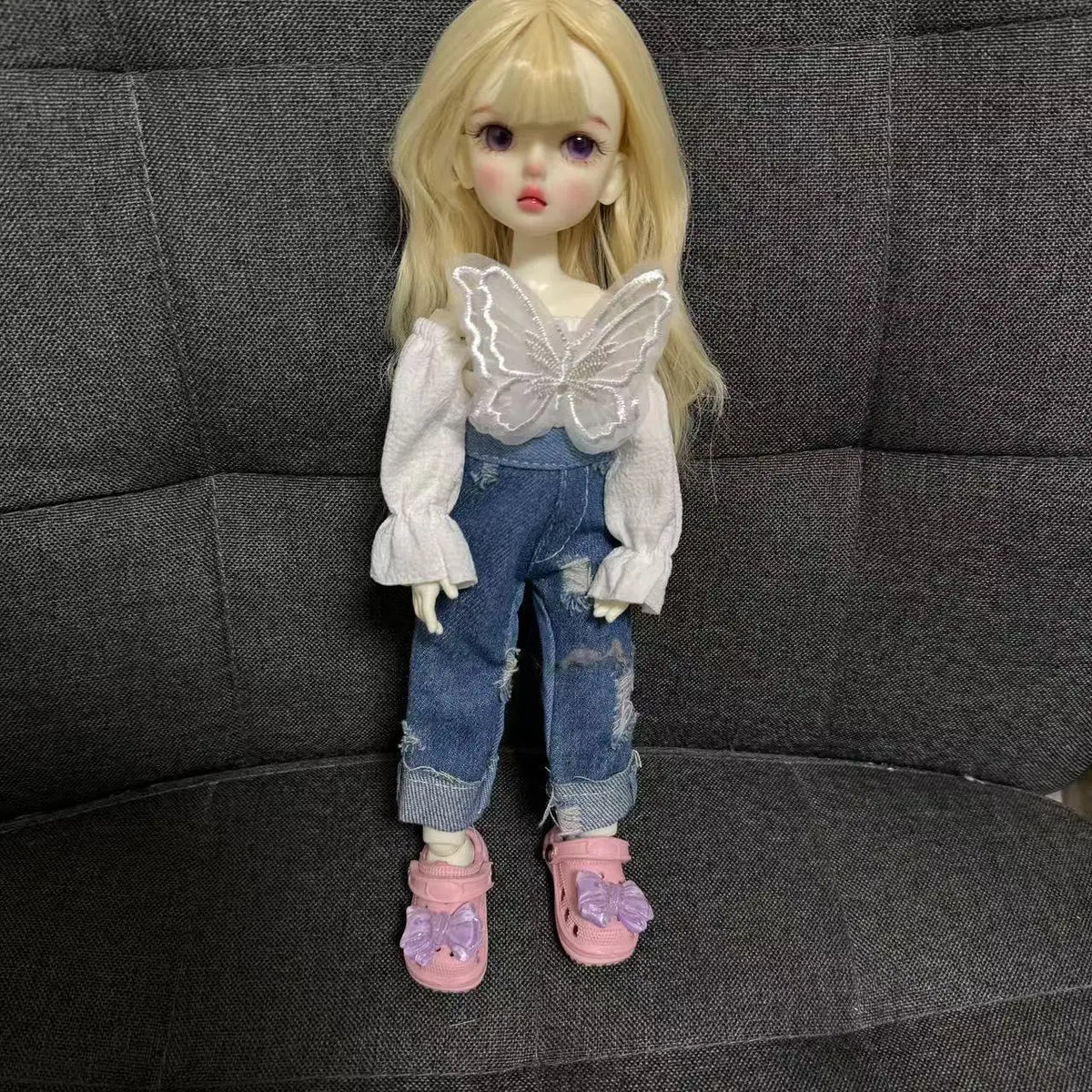 A fashion doll wearing denim overalls and a white shirt with a butterfly detail, standing in front of a dark gray couch.