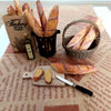 A miniature baguette is displayed alongside a basket of breadsticks and packaged in a brown paper bag.