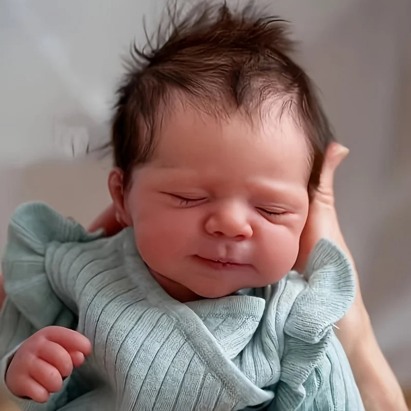 A close-up photo of a newborn Reborn doll with soft cloth body, wearing a light blue knitted garment.