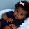 A close-up photo of a reborn baby doll with hand-rooted curly hair wearing a dark blue outfit and a pink bow, resting on whit