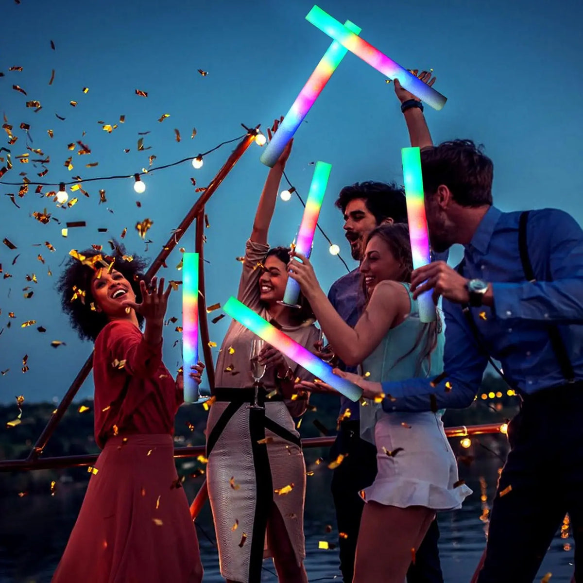 A group of people celebrating outdoors with glowing foam LED glow sticks in the air against a twilight sky.