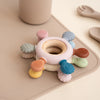 A set of colorful rudder-shaped silicone baby teethers with wooden rings arranged on a light beige tray.