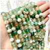 A hand holds a strand of natural stone beads featuring various shades of green, white, and amber stones.