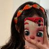A close-up photo of a person wearing an orange and green strawberry crochet headband while holding a smartphone.
