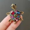 A gold-toned rhinestone turtle brooch pin with colorful gemstone accents is held in a hand.