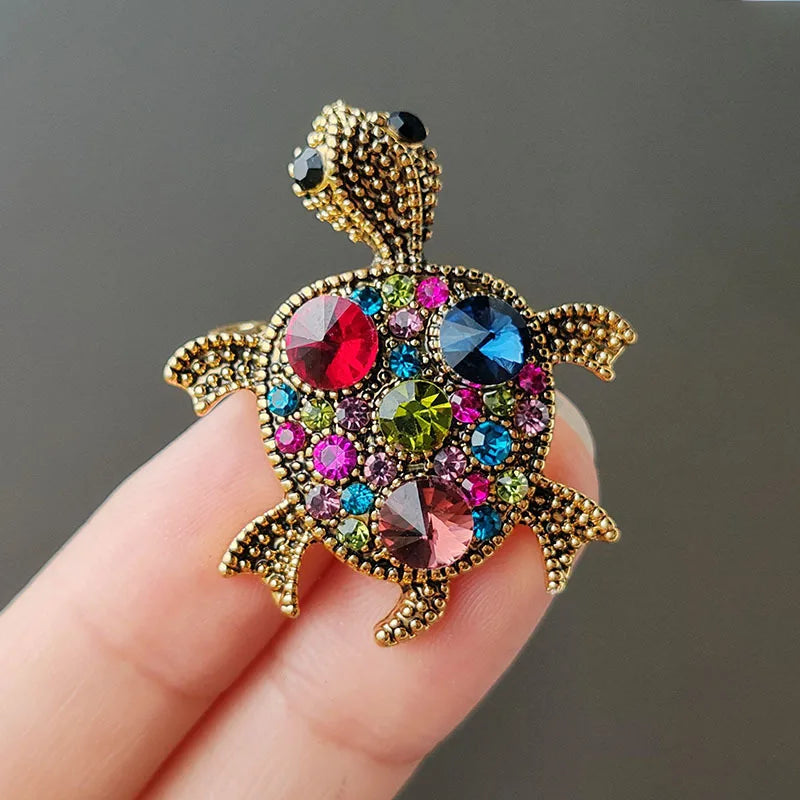 A gold-toned rhinestone turtle brooch pin with colorful gemstone accents is held in a hand.