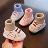 A hand holds a pair of baby soft sole first walker shoes in various pastel colors with bear designs.