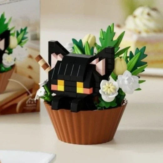 A black cat building block figurine is nestled in a terracotta pot filled with green foliage and white flowers.