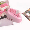 A pink lever action embosser tool is shown next to a bouquet of pink and white flowers.