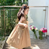 A young girl wearing a beige sleeveless cotton sundress stands outdoors near a glass railing.