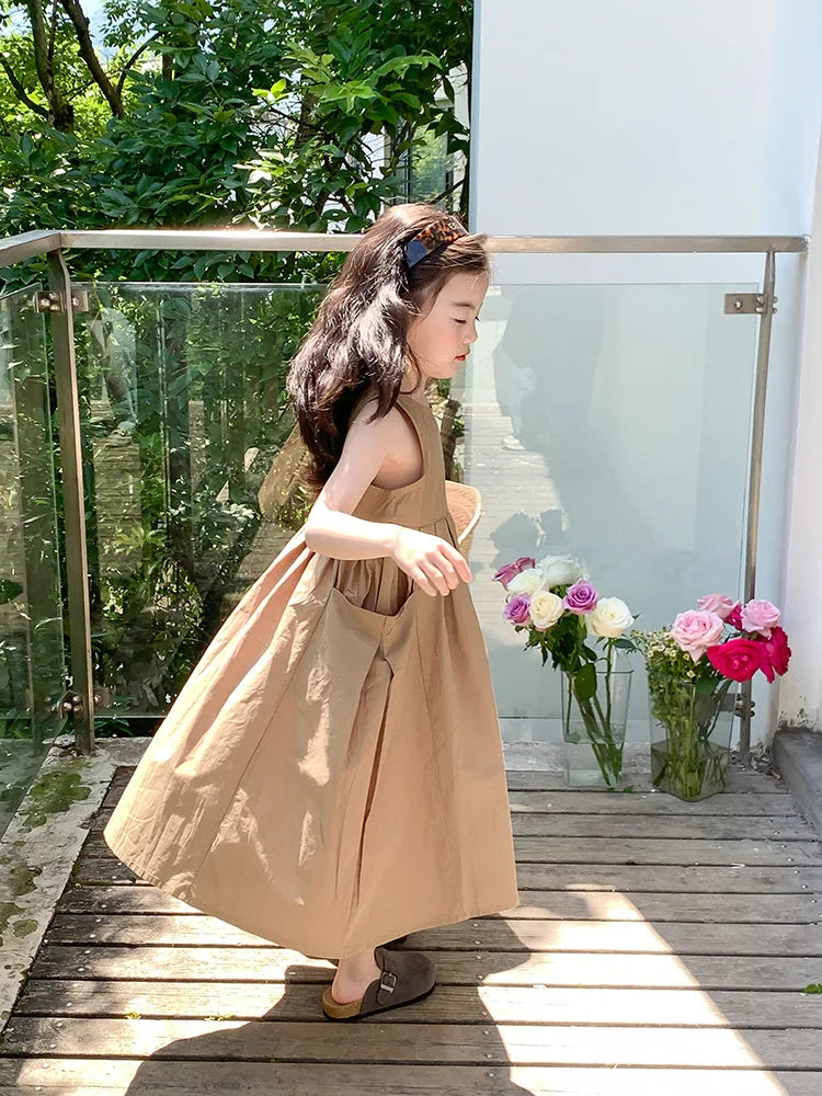 A young girl wearing a beige sleeveless cotton sundress stands outdoors near a glass railing.