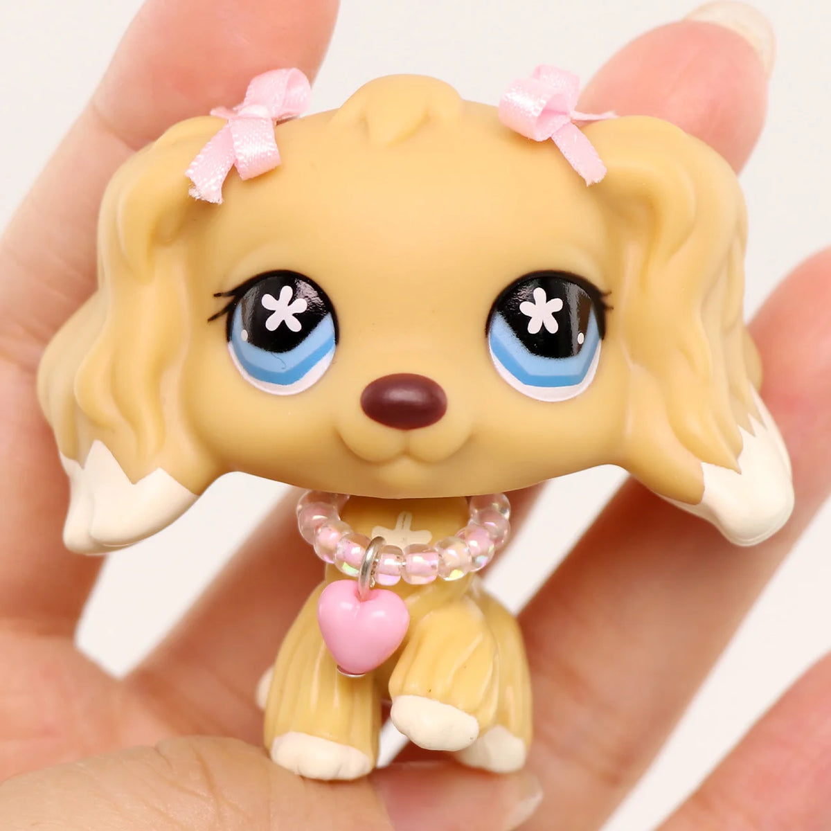 A small, light beige miniature dog figure with pink bows and a pink heart accessory is held in a hand.