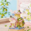 A miniature wooden cabin building set featuring pink and yellow components, flowers, and a windmill on a light blue base.
