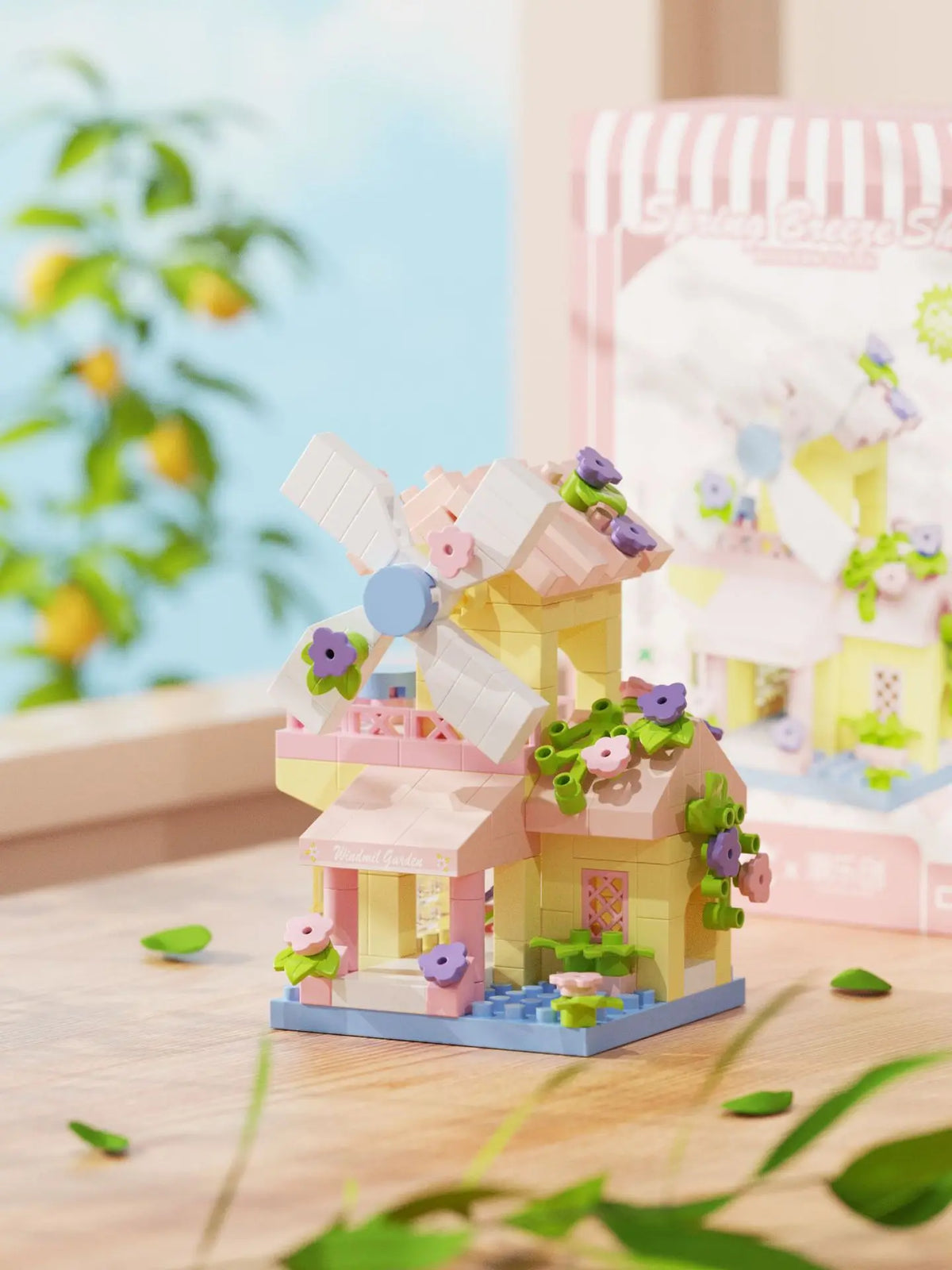 A miniature wooden cabin building set featuring pink and yellow components, flowers, and a windmill on a light blue base.