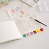 An open watercolor painting book for kids showing line art designs of fruits and animals with colored paint chips.