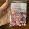 A clear plastic bag contains a variety of pink and purple acrylic loose beads for jewelry making.