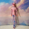 A pink and purple poseable plush doll figure with long purple pants standing against a cloudy sky background.