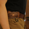 A close-up of a handcrafted brown leather knife sheath with a belt loop worn on a tan fabric, held by a person's arm.