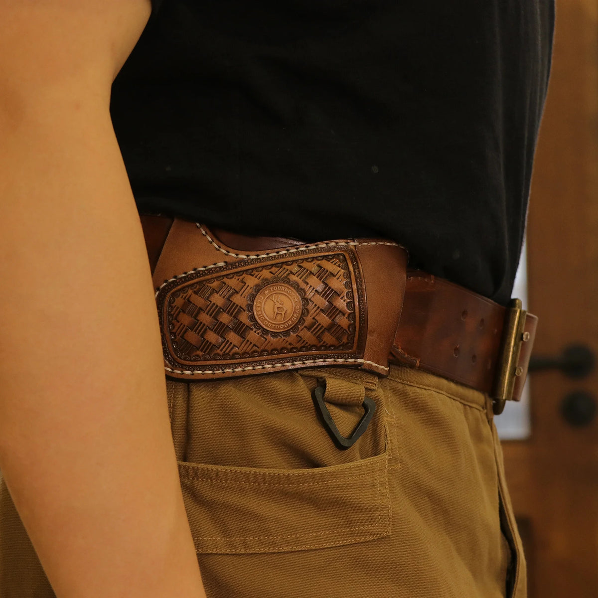 A close-up of a handcrafted brown leather knife sheath with a belt loop worn on a tan fabric, held by a person's arm.