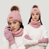 A woman models a pink knit winter hat, scarf, and gloves set with a faux fur pom on a light background.