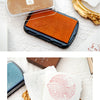 A vintage craft oil-based ink pad in orange-brown color is shown next to white paper and a gold-colored object.