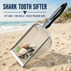 A silver shark tooth sifter tool is shown scooping seashells and small treasures from light-colored beach sand.