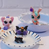Three small figures of animals, including a pink bunny, a black dog wearing purple glasses, and another figure on a white pla