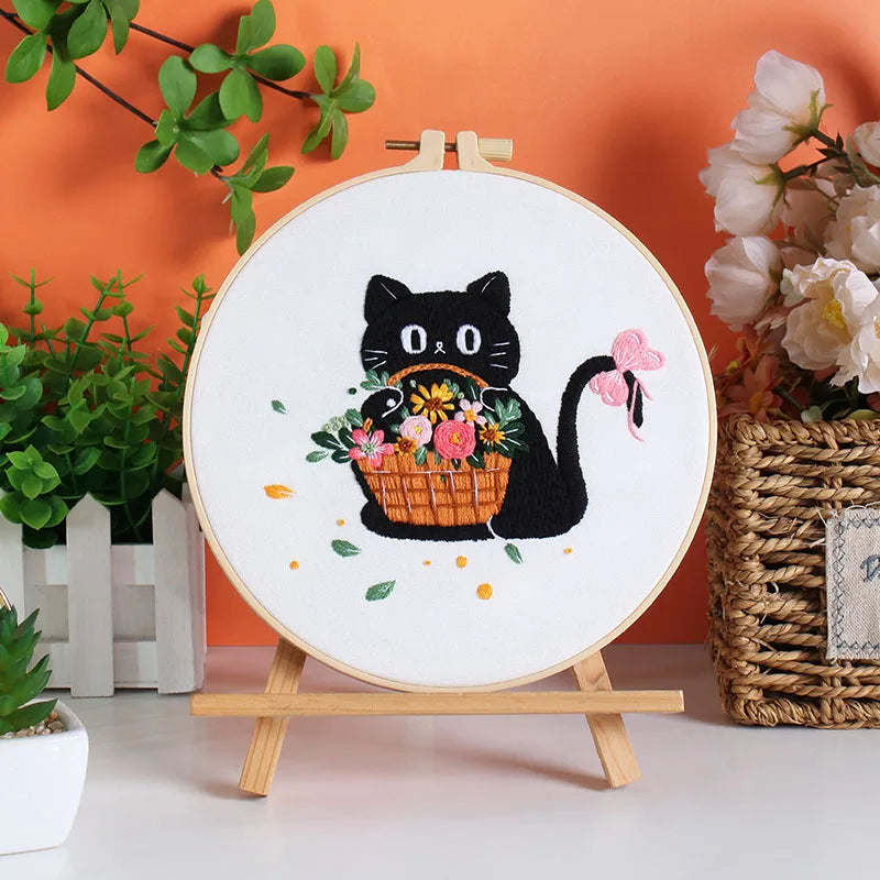 A circular embroidery kit featuring a black cat design on white fabric, displayed on a wooden stand.