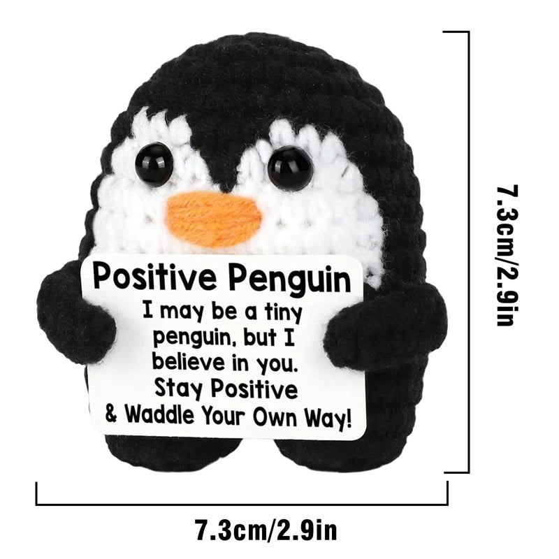 A handmade crochet penguin doll with black and white markings holding a card that reads Stay Positive & Waddle Your Own Way