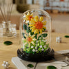A glass dome display of a buildable eternal rose flower kit featuring yellow and orange flowers and green foliage.